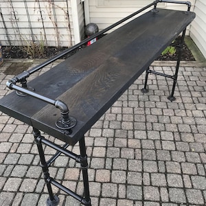 May include: A black wooden bar table with a black metal frame and a black metal handrail. The table is standing on a gray brick patio.