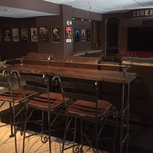 May include: Four black metal bar stools with brown cushions sit in front of a wooden bar counter. The bar counter is made of reclaimed wood and has a metal pipe frame. The bar counter is in front of a brown leather couch.