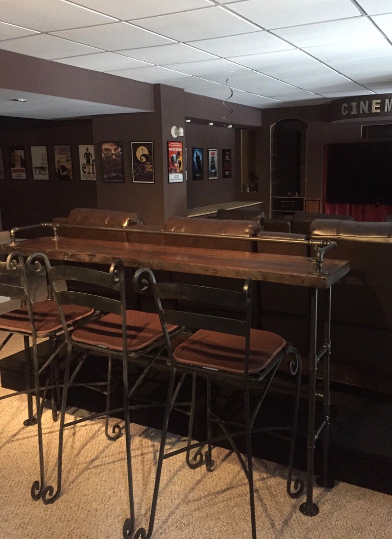 May include: Four black metal bar stools with brown cushions sit in front of a wooden bar counter. The bar counter is made of reclaimed wood and has a metal pipe frame. The bar counter is in front of a brown leather couch.