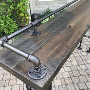 May include: A rustic wooden bar top with a black metal pipe railing. The bar top is stained a dark brown color and has a natural wood grain pattern. The metal railing is attached to the bar top with black metal flanges.