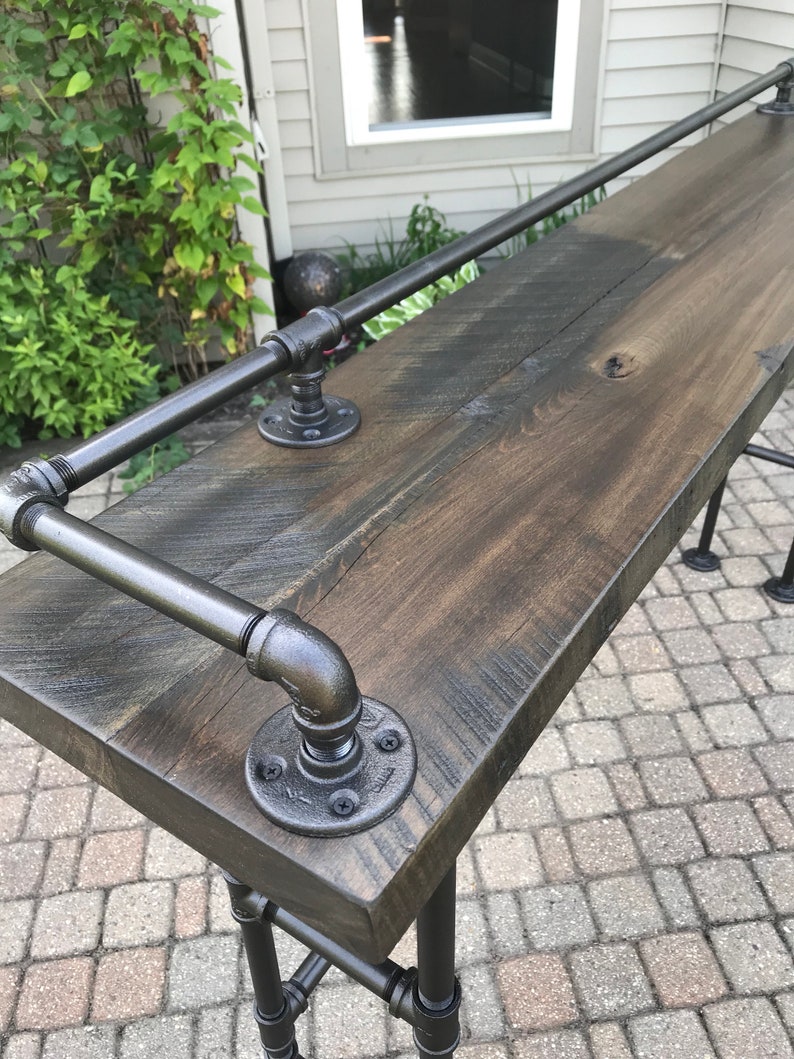 May include: A rustic wooden bar top with a black metal pipe railing. The bar top is stained a dark brown color and has a natural wood grain pattern. The metal railing is attached to the bar top with black metal flanges.