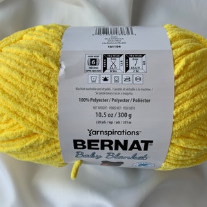 Puede incluir: Un ovillo de hilo amarillo Bernat Baby Blanket. La etiqueta dice "100% Poli&eacute;ster" y "10.5 oz / 300 g".