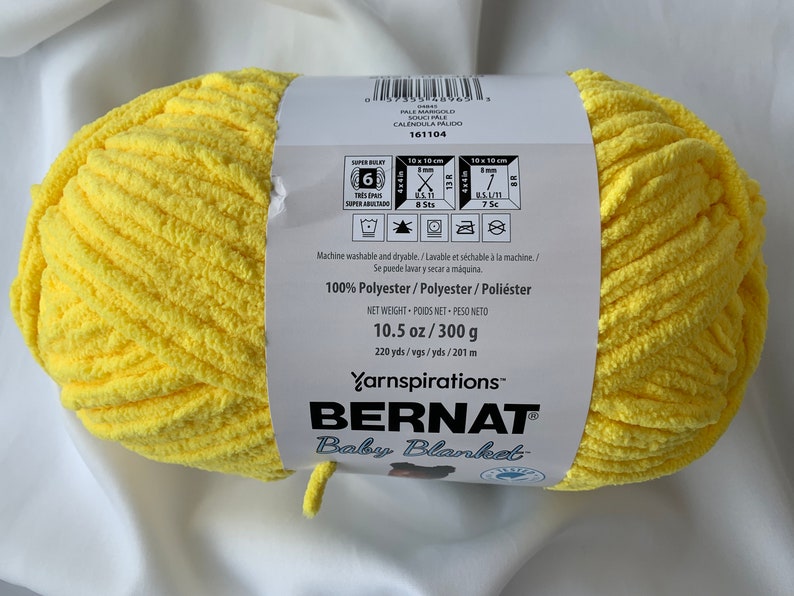 Puede incluir: Un ovillo de hilo amarillo Bernat Baby Blanket. La etiqueta dice "100% Poli&eacute;ster" y "10.5 oz / 300 g".
