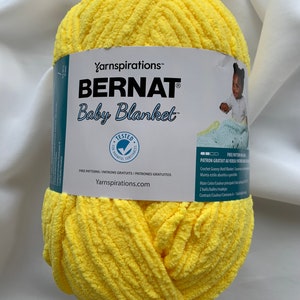 Puede incluir: Una bola de hilo Bernat Baby Blanket de color amarillo. La etiqueta dice "Yarnspirations&trade; BERNAT Baby Blanket&trade;" e incluye instrucciones para una manta de ganchillo con motivo de abuela.