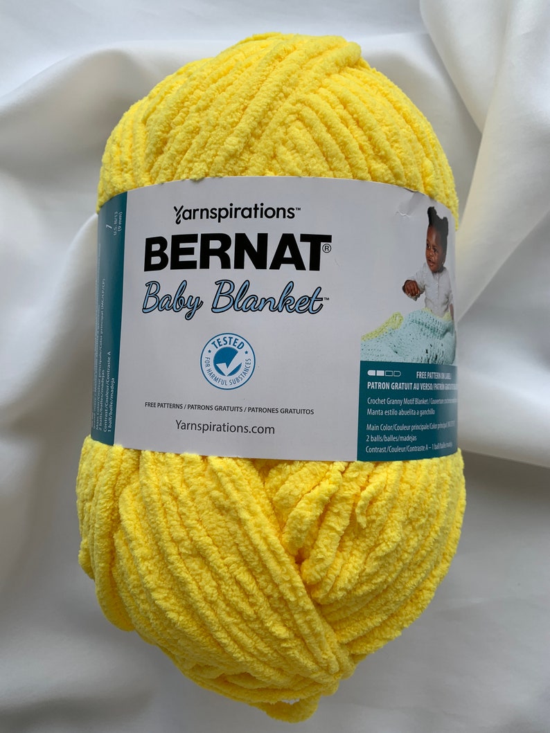 Puede incluir: Una bola de hilo Bernat Baby Blanket de color amarillo. La etiqueta dice "Yarnspirations&trade; BERNAT Baby Blanket&trade;" e incluye instrucciones para una manta de ganchillo con motivo de abuela.