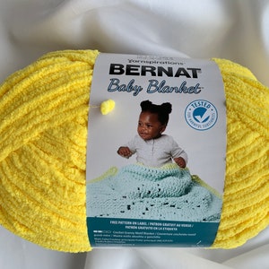 Puede incluir: Una bola de hilo Bernat Baby Blanket de color amarillo con una etiqueta que dice "Bernat Baby Blanket" y tiene una imagen de un beb&eacute; sosteniendo una manta de ganchillo. La etiqueta tambi&eacute;n dice "Free Pattern on Label/Patron Gratuit au Verso/Patron Gratuito en la Etiqueta".
