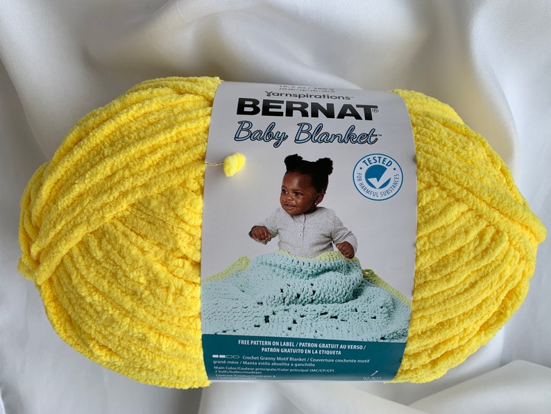 Puede incluir: Una bola de hilo Bernat Baby Blanket de color amarillo con una etiqueta que dice "Bernat Baby Blanket" y tiene una imagen de un beb&eacute; sosteniendo una manta de ganchillo. La etiqueta tambi&eacute;n dice "Free Pattern on Label/Patron Gratuit au Verso/Patron Gratuito en la Etiqueta".