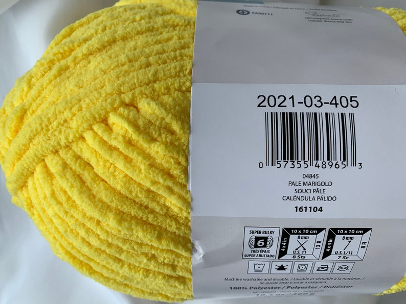 Puede incluir: Una madeja de hilo amarillo super grueso, etiquetada como "Pale Marigold" con el c&oacute;digo de producto "2021-03-405". El hilo es 100% poli&eacute;ster y se puede lavar y secar a m&aacute;quina.