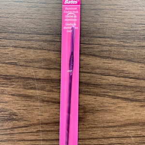 May include: A purple aluminum crochet hook packaged on a pink card with the brand name "Susan Bates" and the text "Aluminum Crochet Hook" in English and Spanish.
