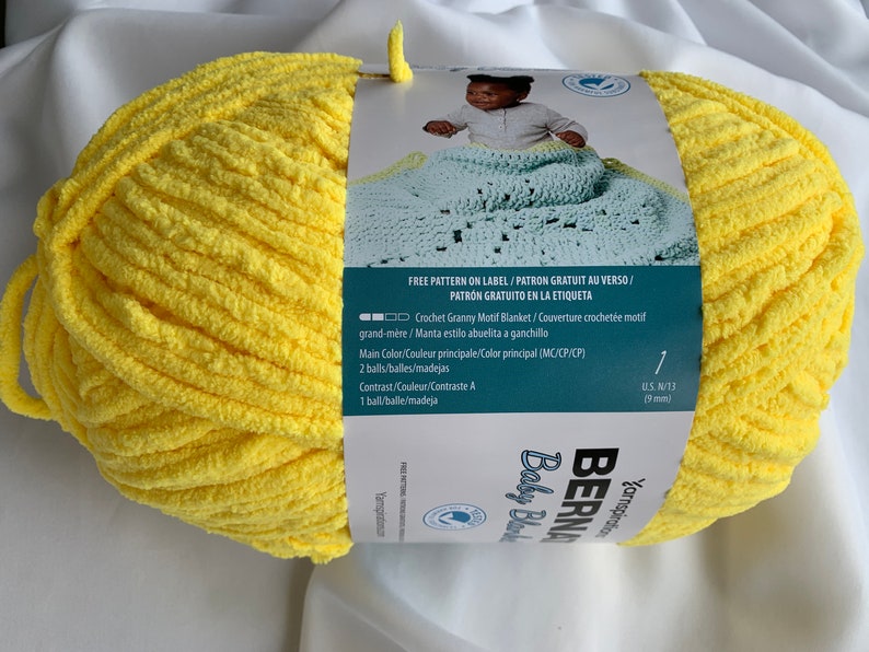 Puede incluir: Una madeja de hilo amarillo Bernat Baby Blanket con una etiqueta que dice "Free Pattern on Label/Patron Gratuit au Verso/Patron Gratuito en la Etiqueta" e incluye instrucciones para tejer una manta de beb&eacute; con motivo de granny. La etiqueta tambi&eacute;n dice "Crochet Granny Motif Blanket/Couverture crochet&eacute;e motif grand-m&egrave;re/Manta estilo abuelita a ganchille".
