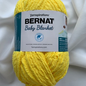 Puede incluir: Una bola de hilo Bernat Baby Blanket de color amarillo. La etiqueta dice "Yarnspirations&trade; Bernat Baby Blanket" e incluye el texto "Free Patterns/Patrons Gratuits/Patrones Gratuitos" y "Yarnspirations.com".