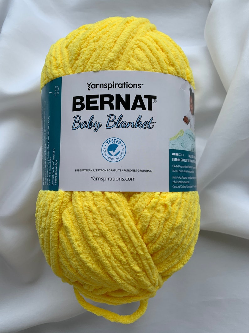Puede incluir: Una bola de hilo Bernat Baby Blanket de color amarillo. La etiqueta dice "Yarnspirations&trade; Bernat Baby Blanket" e incluye el texto "Free Patterns/Patrons Gratuits/Patrones Gratuitos" y "Yarnspirations.com".