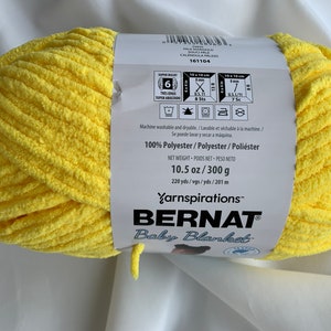 Puede incluir: Una bola de hilo Bernat Baby Blanket de color amarillo. La etiqueta dice "100% Poli&eacute;ster" y "10.5 oz/300 g".