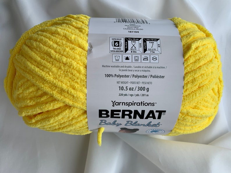 Puede incluir: Una bola de hilo Bernat Baby Blanket de color amarillo. La etiqueta dice "100% Poli&eacute;ster" y "10.5 oz/300 g".