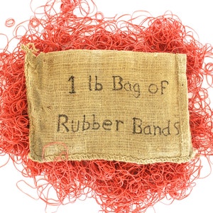May include: A burlap bag labeled "1 lb Bag of Rubber Bands" is surrounded by a pile of red rubber bands. The bag is tan with black lettering. The rubber bands are scattered around the bag on a white background.