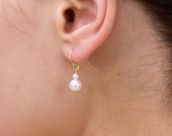 May include: A close-up of a person's ear wearing a pair of gold drop earrings. Each earring features a small, round pearl suspended below a larger pearl. The earrings are simple and elegant.