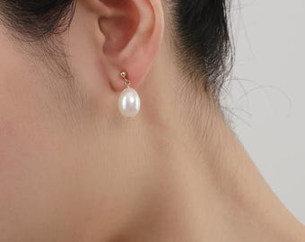 May include: A close-up of a person wearing a gold stud earring with a white teardrop-shaped pearl. The earring is a classic design, suitable for everyday wear or special occasions. The pearl has a smooth, lustrous surface.