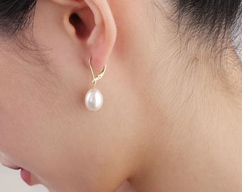 May include: A gold-colored earring with a white, teardrop-shaped pearl. The earring is attached to the ear with a lever-back clasp. The pearl hangs freely, creating a classic and elegant look. The earring is shown on a person's ear.