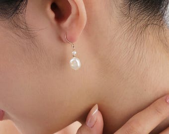 May include: A gold-colored earring with a small, round pearl above a larger, teardrop-shaped pearl. The earring is hanging from a person's ear. The person's skin is fair, and their hair is dark.
