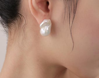 May include: A close-up of a white, baroque pearl stud earring. The earring has an irregular, organic shape and a lustrous surface. The earring is worn on the ear of a person.