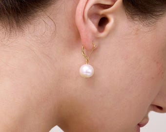 May include: A gold-colored earring with a large, round, white pearl hangs from a woman's ear. The earring is a leverback style. The woman's skin is fair, and her hair is dark. The background is a soft, neutral color.