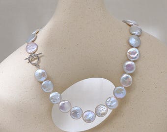 May include: A pearl necklace with large, flat, iridescent pearls. The necklace is fastened with a silver toggle clasp. The pearls have a soft, pearlescent sheen, and the necklace is displayed on a cream-colored vase.