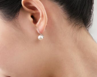 May include: A close-up of a single pearl earring. The earring features a small, white pearl suspended from a silver hook. The hook has a curved, swirling design. The earring is worn on a person's ear.