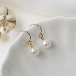 Op de afbeelding: Een paar goudkleurige oorhangers met witte, ronde parels. De oorbellen worden gepresenteerd op een wit, gebogen oppervlak. De tekst "PearlFans" is zichtbaar onderaan de afbeelding.