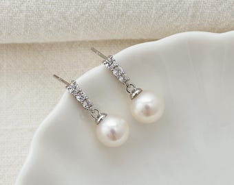 May include: A pair of silver-toned drop earrings with a pearl and rhinestone design. Each earring features a row of clear rhinestones and a dangling white pearl. The earrings are displayed on a white, scalloped plate. The text "PearlFans" is visible.