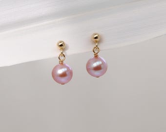 May include: A pair of gold-colored stud earrings with dangling, round, light pink pearls. The earrings are displayed against a soft, neutral background with a white, textured element in the upper left corner.