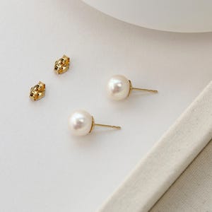 Op de afbeelding: Een paar parelstud-oorbellen met gouden pinnen en twee gouden oorbelruggen. De oorbellen zijn geplaatst tegen een witte achtergrond met een neutraal gekleurde stof.