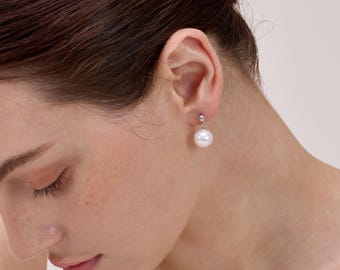 May include: A pair of pearl drop earrings. Each earring features a large, round, white pearl suspended from a small, silver-colored stud. The earrings are shown on a person's ear, with the focus on the jewelry.