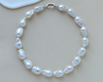 May include: A necklace featuring large, irregularly shaped white pearls. The pearls are strung together with a silver-colored clasp. The necklace is displayed on a neutral-colored surface, with a white, organic-shaped element in the background.