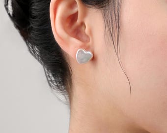 May include: A close-up of a person's ear with a heart-shaped earring. The earring is a light, pearly color and has a smooth, slightly irregular surface. The earring is a stud style, and the image is well-lit.