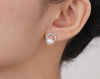 May include: A pearl stud earring with a halo of small, clear crystals. The earring is set in a silver-colored metal and is worn on the ear. The pearl is round and white, and the crystals sparkle.