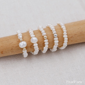 Puede incluir: Cinco delicados anillos de perlas con detalles dorados. Cada anillo está hecho de pequeñas perlas blancas enhebradas en un fino alambre dorado. Los anillos están apilados uno encima del otro sobre una superficie de madera.