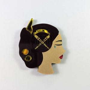 May include: A brown and gold resin brooch depicting a woman's profile with a tear on her cheek. The woman has a gold headband with a cross design and two gold circles on her ear.