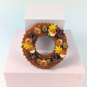 May include: A brown wooden wreath decorated with fall-themed elements. The wreath features pumpkins, acorns, pinecones, and colorful leaves in shades of orange, yellow, and burgundy. The wreath is displayed on a white and pink surface.
