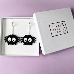 May include: A pair of black spider earrings with white eyes and silver chain attachments are displayed inside a white box. The box has the text "chikai shop .com" printed on the lid in a brown square.