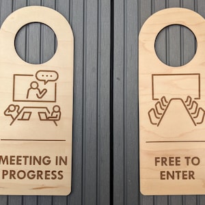 Puede incluir: Dos colgadores de puerta de madera con un agujero en la parte superior para colgar en una puerta. El colgador de la izquierda tiene un dibujo de una reunión en curso con una persona presentando en una pantalla y dos personas en portátiles. El texto en el colgador de la izquierda dice "Reunión en curso". El colgador de la derecha tiene un dibujo de un cine con filas de asientos y el texto "Entrada libre".