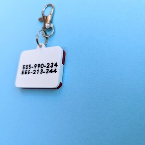 May include: White plastic key chain with a red border and two phone numbers printed on it: 555-990-234 and 555-213-344.