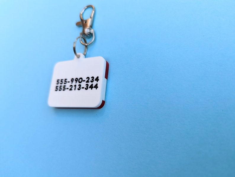 May include: White plastic key chain with a red border and two phone numbers printed on it: 555-990-234 and 555-213-344.