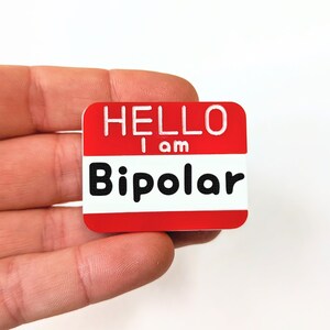 May include: Red name tag with white text that reads "Hello I am Bipolar".