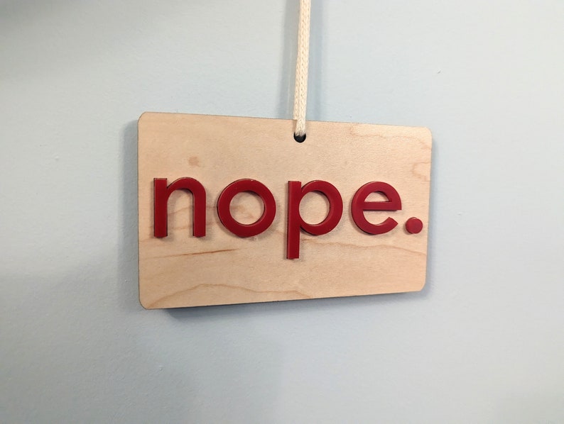 Puede incluir: Un letrero de madera con la palabra "nope" en letras rojas. El letrero est&aacute; colgado de una cuerda blanca.