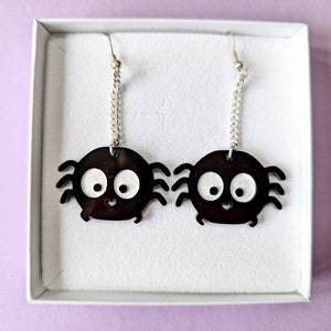 May include: A pair of black spider earrings with white eyes and a smiling mouth. The earrings hang from silver-colored chains and hooks, presented in a white box against a light purple background.