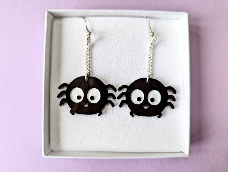May include: A pair of black spider earrings with white eyes and a smiling mouth. The earrings hang from silver-colored chains and hooks, presented in a white box against a light purple background.