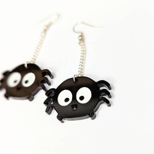 May include: A pair of black spider earrings with white eyes and silver chain accents. The earrings are made of a glossy material and are designed with a cartoonish spider shape. The earrings are on a white background.