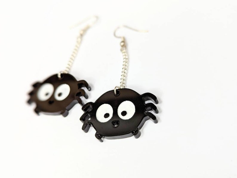 May include: A pair of black spider earrings with white eyes and silver chain accents. The earrings are made of a glossy material and are designed with a cartoonish spider shape. The earrings are on a white background.