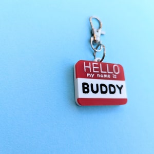 May include: Red and white plastic name tag with a silver clip. The tag says "HELLO my name is BUDDY".