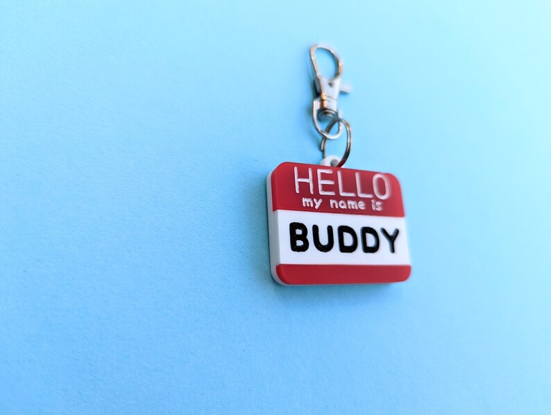May include: Red and white plastic name tag with a silver clip. The tag says "HELLO my name is BUDDY".